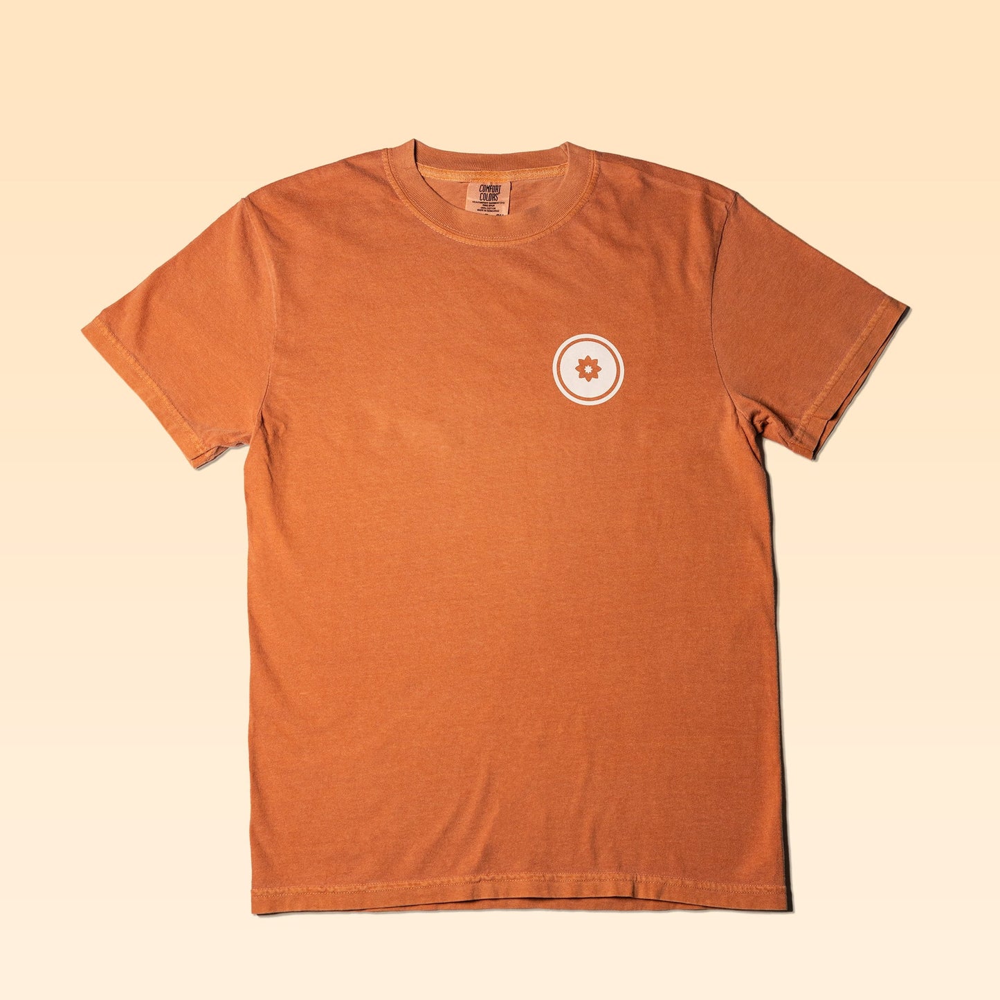 An orange Worth The Trip Shirt by Mescalito Coffee with a small circular white gear emblem on the left chest, displayed flat on a light beige background.