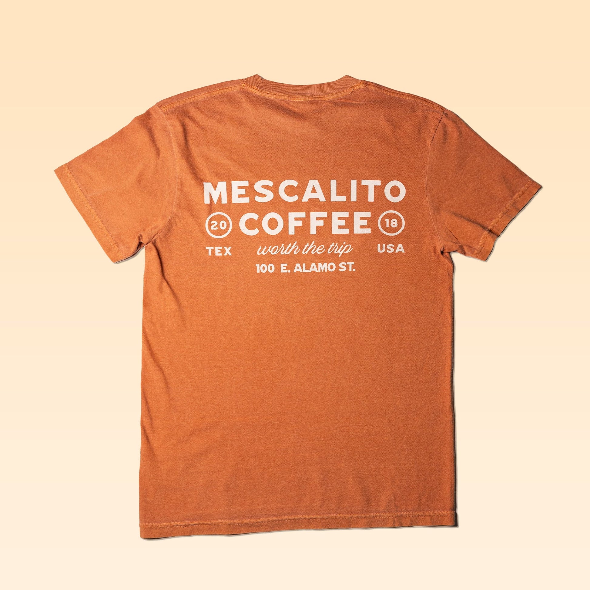 The Worth The Trip Shirt by Mescalito Coffee features rust fabric, white back text ("Mescalito Coffee, 2018, Tex USA, worth the trip, 100 E. Alamo St.")—a must-have for Brenham coffee fans and scenic explorers.