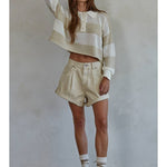 A woman stands against a plain wall, wearing a beige striped knit sweater, socks, brown sandals, and By Together’s Woven Cotton Denim Shorts that exude vintage charm.