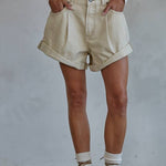 A person wears By Together’s Woven Cotton Denim Shorts in light beige with a cropped sweater, socks, and strappy sandals, standing hands in pockets against a plain background and radiating vintage charm.