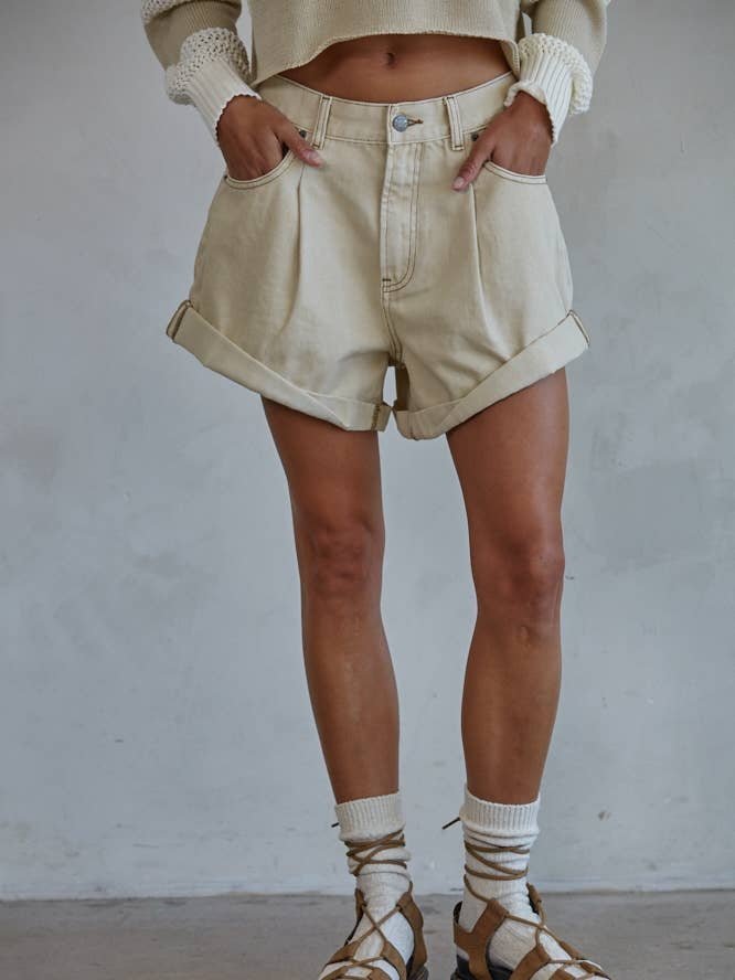 A person wears By Together’s Woven Cotton Denim Shorts in light beige with a cropped sweater, socks, and strappy sandals, standing hands in pockets against a plain background and radiating vintage charm.