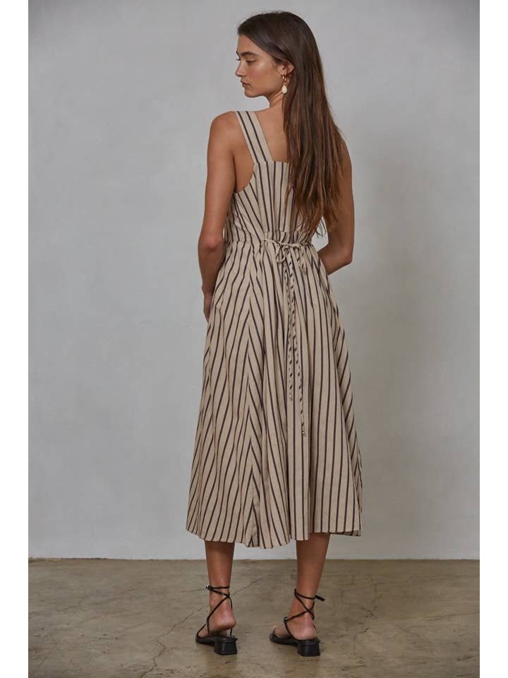 Woman stands on a concrete floor facing away, wearing By Together's Woven Cotton Striped Square Neck Flare Midi Dress with vertical black stripes and a waist tie, paired with black sandals.