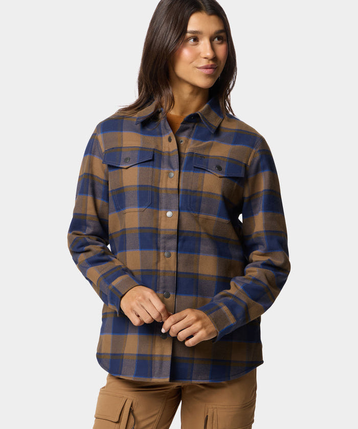 A woman wears the Duck Camp W's Campfire Shacket Pintail Plaid, featuring blue and brown plaid with chest pockets, paired with tan cargo pants, standing against a plain light background.