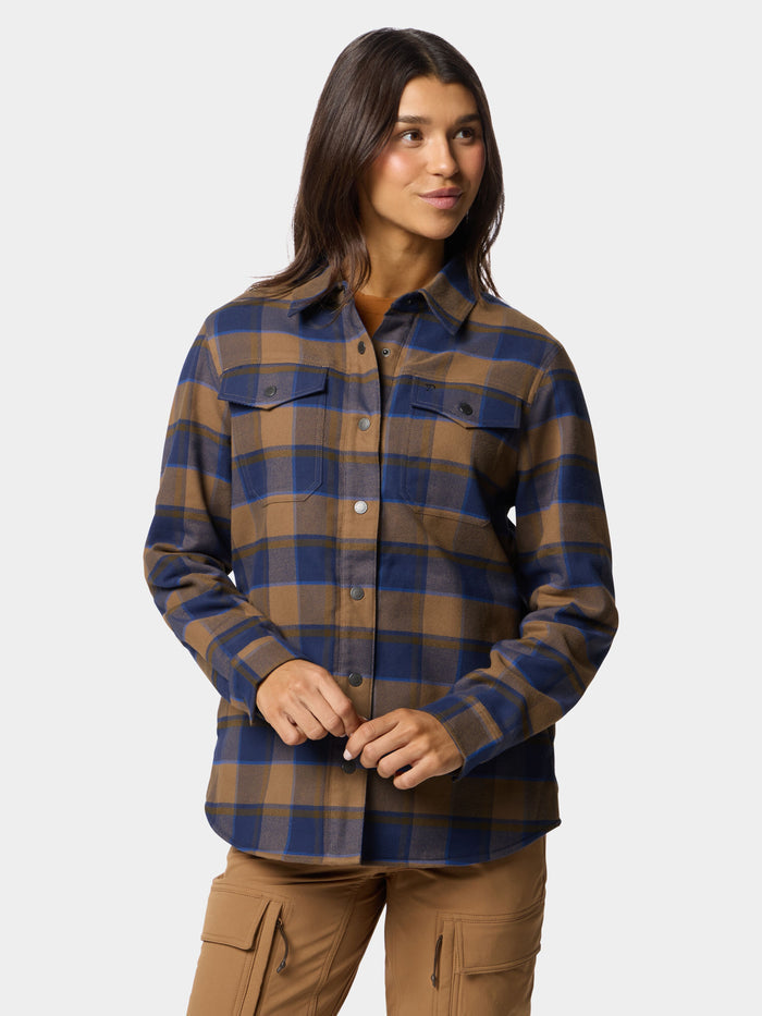 A woman wears the Duck Camp W's Campfire Shacket Pintail Plaid, featuring blue and brown plaid with chest pockets, paired with tan cargo pants, standing against a plain light background.