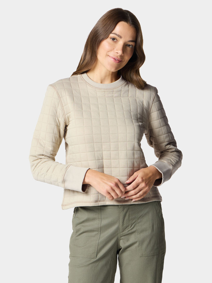 A person stands against a plain background wearing Duck Camp's W's Cheyenne Quilted Crew in light beige—an ideal layering piece—paired with olive green pants.