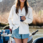 A woman in Duck Camp’s W's Lightweight Fishing Shirt Long Sleeve smiles by a river, wearing sunglasses and a hat while holding a canned drink.