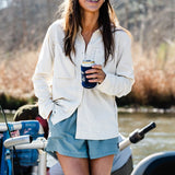 A woman in Duck Camp’s W's Lightweight Fishing Shirt Long Sleeve smiles by a river, wearing sunglasses and a hat while holding a canned drink.