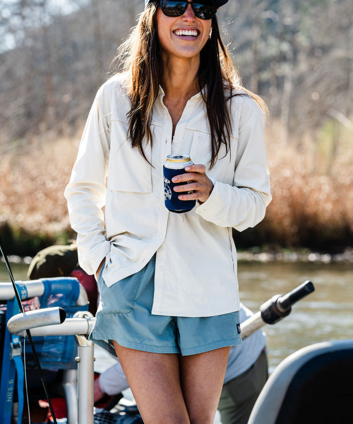 A woman in the Duck Camp W's Lightweight Fishing Shirt Long Sleeve, sunglasses, a cap, and shorts stands outdoors holding a canned drink with a blue koozie.
