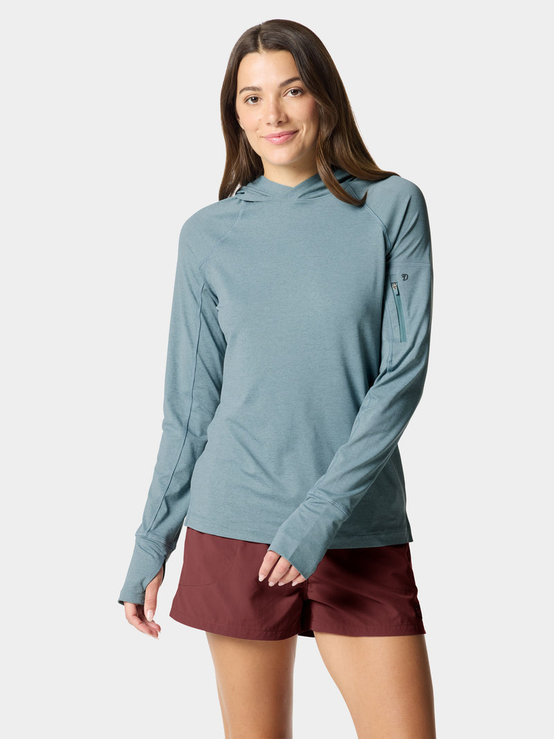 A woman models the Duck Camp W's Rockport Hoodie in Heathered Trooper, a cooling, teal long-sleeve with a zippered sleeve pocket, paired with maroon shorts against a plain light background.