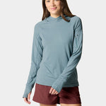 A woman models the Duck Camp W's Rockport Hoodie in Heathered Trooper, a cooling, teal long-sleeve with a zippered sleeve pocket, paired with maroon shorts against a plain light background.