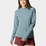 A woman models the Duck Camp W's Rockport Hoodie in Heathered Trooper, a cooling, teal long-sleeve with a zippered sleeve pocket, paired with maroon shorts against a plain light background.