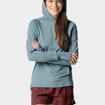 A woman smiles with arms crossed, wearing the Duck Camp W's Rockport Hoodie Heathered Trooper—a blue UPF 50+ cooling long-sleeve shirt—paired with maroon shorts and a blue cap featuring a fish graphic.
