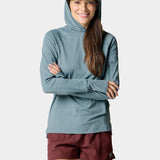 A woman smiles with arms crossed, wearing the Duck Camp W's Rockport Hoodie Heathered Trooper—a blue UPF 50+ cooling long-sleeve shirt—paired with maroon shorts and a blue cap featuring a fish graphic.