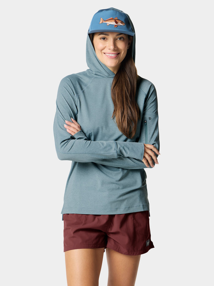 A woman smiles with arms crossed, wearing the Duck Camp W's Rockport Hoodie Heathered Trooper—a blue UPF 50+ cooling long-sleeve shirt—paired with maroon shorts and a blue cap featuring a fish graphic.