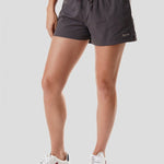 A person wearing a burgundy shirt, Duck Camp W's Scout Shorts in dark gray, and white sneakers stands against a plain light background.