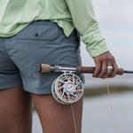 A person in a light green shirt and Duck Camp W's Scout Shorts holds a fly fishing rod and reel by their side, standing near water.