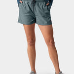A person stands with hands in pockets wearing a blue long-sleeve shirt, Duck Camp W's Scout Shorts with durable nylon and pockets, and gray sneakers against a plain background.