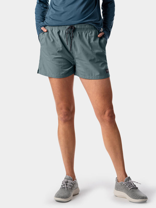A person stands with hands in pockets, wearing a blue long-sleeve shirt, gray sneakers, and Duck Camp's W's Scout Shorts—crafted from lightweight nylon and featuring pockets—against a plain background. Ideal for warm weather.