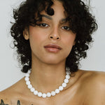 A person with short curly hair and butterfly tattoos poses against a plain background, wearing the Kara Yoo Yona Collar necklace—a handmade statement piece from Vancouver.