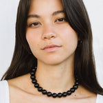 A woman with long dark hair wears a white top and the Yona Collar by Kara Yoo, looking directly at the camera against a plain background.