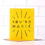 The "You're Magic" greeting card by Daydream Prints features black text with peace, triangle, and wave symbols on yellow card stock. It stands upright on a recycled envelope and is proudly made in Portland.