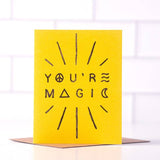 The "You're Magic" greeting card by Daydream Prints features black text with peace, triangle, and wave symbols on yellow card stock. It stands upright on a recycled envelope and is proudly made in Portland.