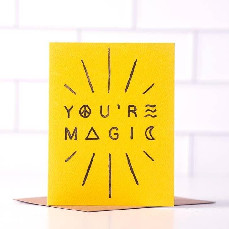 The "You're Magic" greeting card by Daydream Prints features black text with peace, triangle, and wave symbols on yellow card stock. It stands upright on a recycled envelope and is proudly made in Portland.