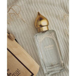 The Amber Eau de Parfum Spray 50ml by Nemat, in a clear glass bottle with a gold cap labeled "Amber," sits on textured beige fabric beside a brown paper bag—an elegant, signature scent in eco-friendly packaging.