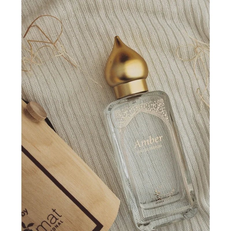 A clear glass bottle of Nemat Amber Eau de Parfum Spray 50ml with a gold cap sits on textured beige fabric, capturing the warm scent of amber, beside a partially visible brown paper bag.