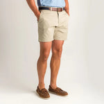 A person wearing Duck Head 7in Gold School shorts, a brown belt, light blue shirt, and brown boat shoes stands with hands in pockets against a plain background.