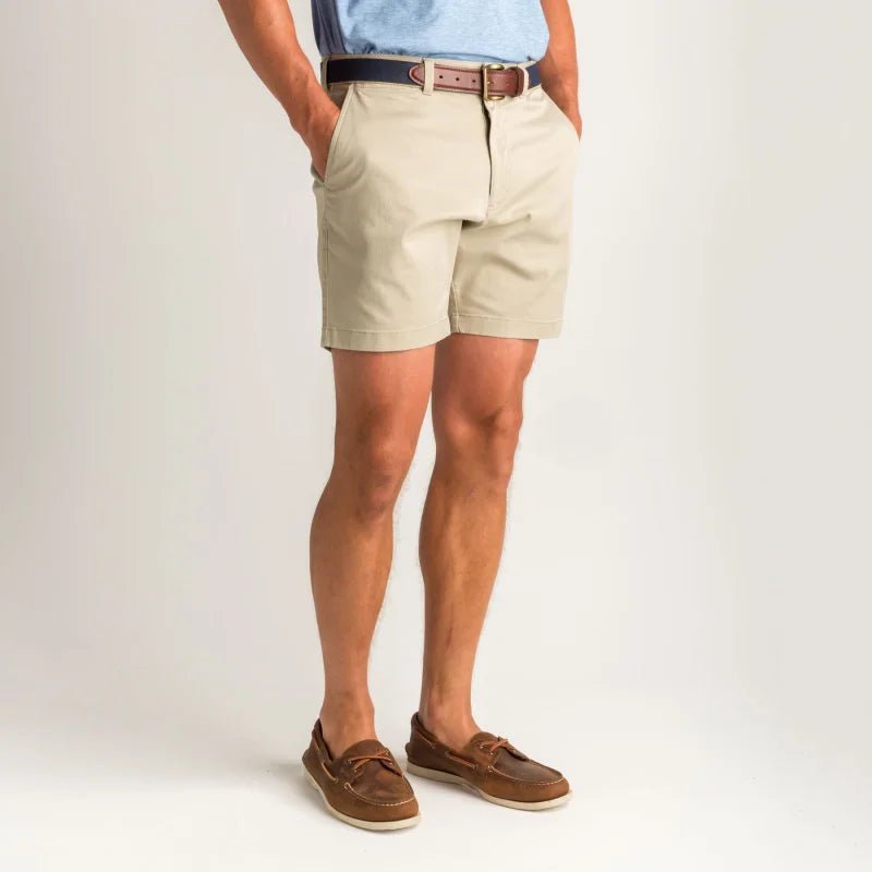 A person wearing Duck Head 7 in Gold School beige chino shorts, a brown belt, light blue shirt, and brown boat shoes stands against a plain light background with hands in pockets.