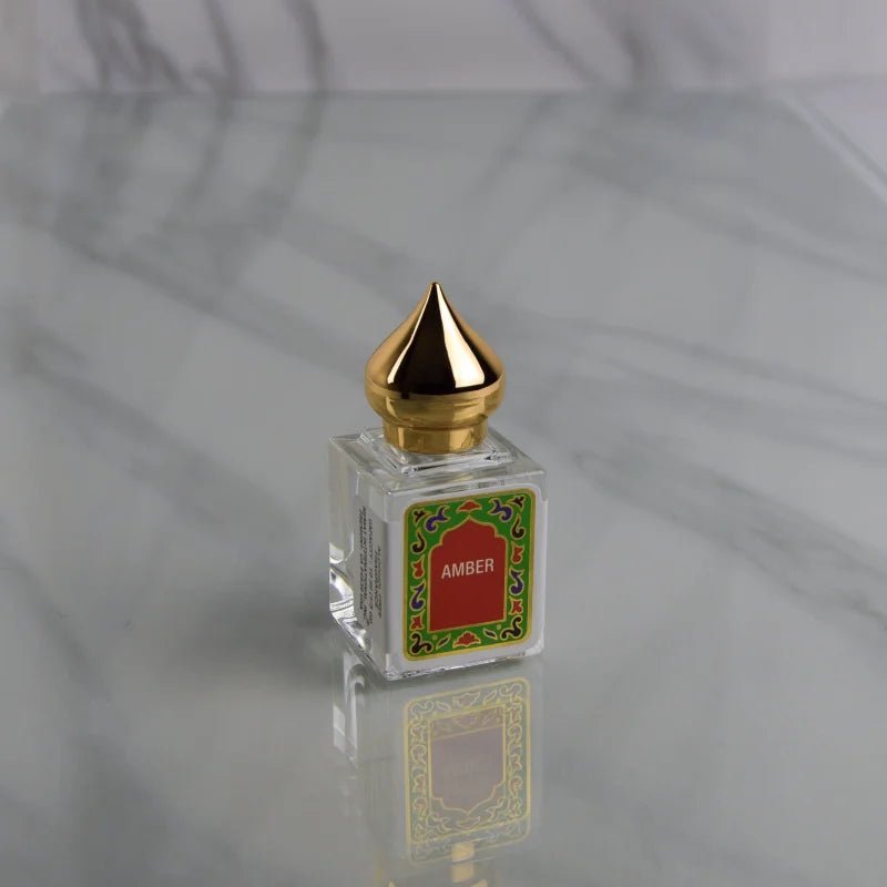 A glass Amber Perfume Oil bottle by Nemat, featuring a gold pointed cap and a decorative "Amber" label, sits on a reflective marble surface, capturing the allure of this signature Nemat fragrance.
