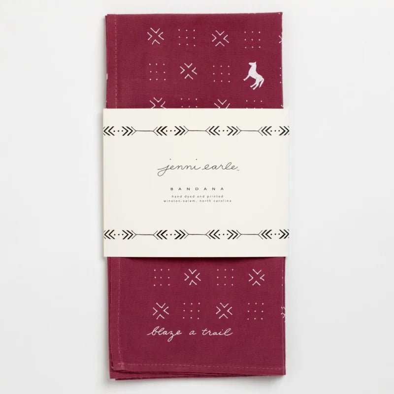The Jenni Earle "Blaze A Trail" bandana is hand-dyed maroon with white geometric patterns, a small fox illustration, and the phrase "Blaze a Trail," wrapped in a branded paper label.