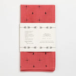 The “Bandana | Explore More” by Jenni Earle is a folded, hand-dyed red cotton bandana with small black geometric and animal motifs, wrapped in a white paper label featuring text and arrow graphics.