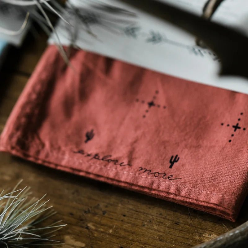 The Jenni Earle Bandana | Explore More, featuring small cactus and cross designs with “explore more” embroidered on the edge, rests on a wooden surface adorned with pine needles.