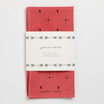 A red cotton bandana, hand-dyed with black cactus and dot patterns, folded and wrapped in a white “Jenni Earle” label reading “Explore More.” Product: Bandana | Explore More | Jenni Earle by Jenni Earle.