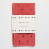 A red cotton bandana, hand-dyed with black cactus and dot patterns, folded and wrapped in a white “Jenni Earle” label reading “Explore More.” Product: Bandana | Explore More | Jenni Earle by Jenni Earle.