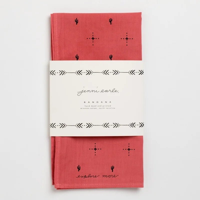 A red cotton bandana, hand-dyed with black cactus and dot patterns, folded and wrapped in a white “Jenni Earle” label reading “Explore More.” Product: Bandana | Explore More | Jenni Earle by Jenni Earle.
