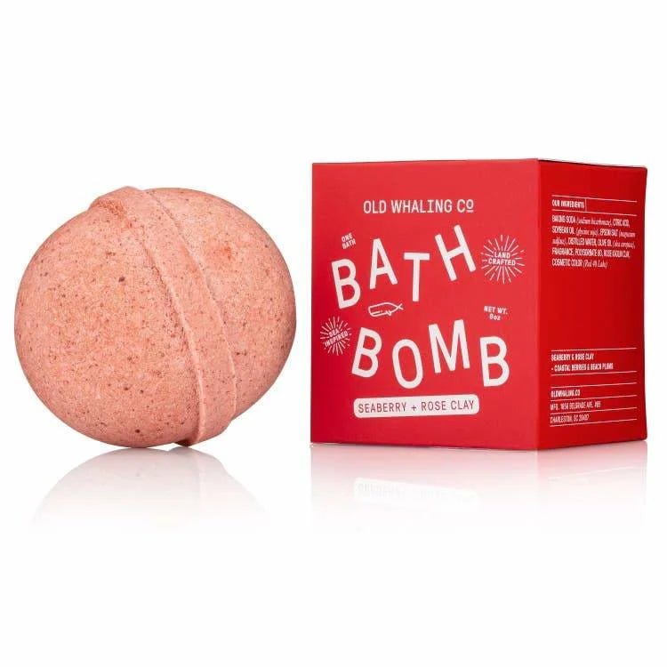 A round pink bath bomb with rose kaolin clay sits beside a red box labeled “Bath Bomb | Seaberry + Rose Clay | Old Whaling Co.” The box lists ingredients like epsom salts on the side.