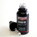 An open bottle of Beard Oil | Uppercut Deluxe, a conditioning beard oil from Uppercut Deluxe with patchouli and leather scent, sits next to its dropper on a white surface.