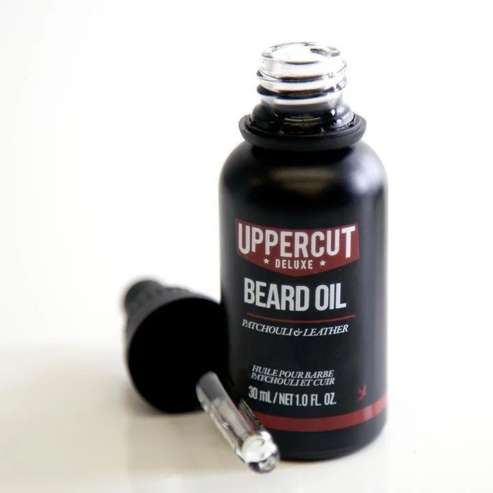 An open bottle of Beard Oil | Uppercut Deluxe, a conditioning beard oil from Uppercut Deluxe with patchouli and leather scent, sits next to its dropper on a white surface.
