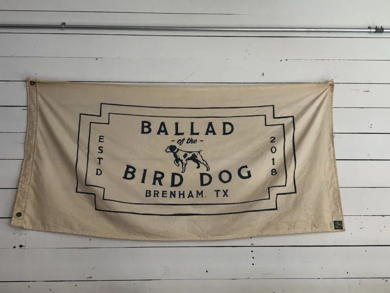 The Bird Dog Flag | Large by Ballad of the Bird Dog features a beige, neon-style design with black "Ballad of the Bird Dog, Brenham, TX, Estd 2018" text and a central dog illustration.