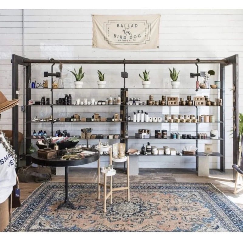 A shop display features shelves with candles, jars, and potted plants, a round table with small items, a rug on the floor, and a glowing neon sign above a large Bird Dog Flag by Ballad of the Bird Dog.