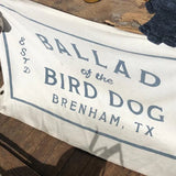 The Bird Dog Flag | Medium by Ballad of the Bird Dog is a white shop flag with a stamp logo reading "Ballad of the Bird Dog, Brenham, TX, Estd 2018," resting on wood with part of a dog's head visible on the left.