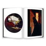 An open book from Anthology Editions’ "Jane Dickson in Times Square" shows cars by a Rocky III sign and a figure facing neon "HOT" and "PARKING" signs—iconic New York City scenes by Jane Dickson.