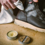A person polishes a leather boot with a cloth, using an open tin of Boot Wax by Otter Wax that's placed on the floor nearby.