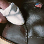 A hand uses a white cloth to polish a brown leather boot with an American flag patch, applying Otter Wax Boot Wax for shine and protection.