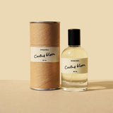 A glass bottle of Cactus Bloom Eau de Parfum by Saguara Perfumes, capped in black, sits beside its cylindrical box on a beige surface, reflecting a scent that's handmade in California.