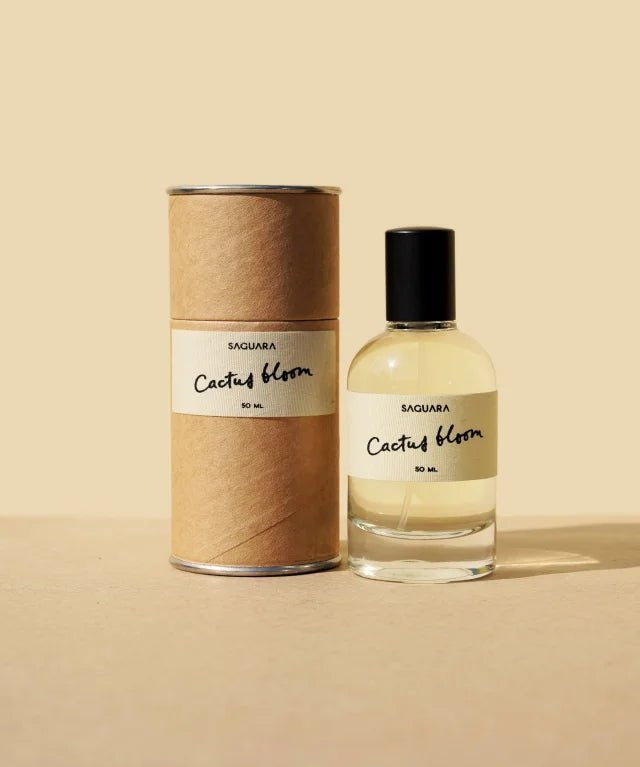 A glass bottle of Cactus Bloom Eau de Parfum by Saguara Perfumes, capped in black, sits beside its cylindrical box on a beige surface, reflecting a scent that's handmade in California.