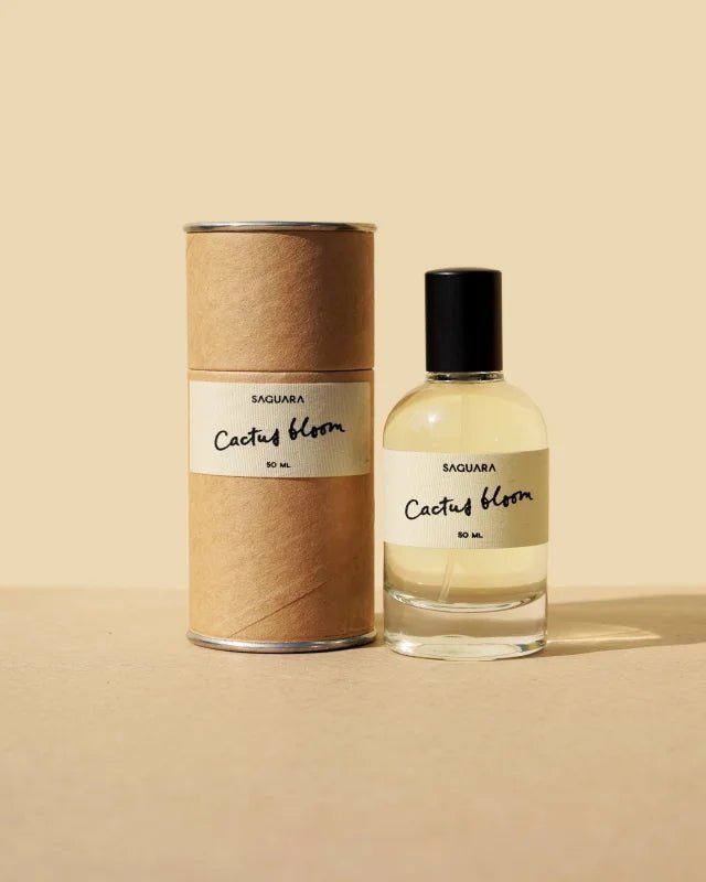 A glass bottle of Cactus Bloom Eau de Parfum by Saguara Perfumes, capped in black, sits beside its cylindrical box on a beige surface, reflecting a scent that's handmade in California.